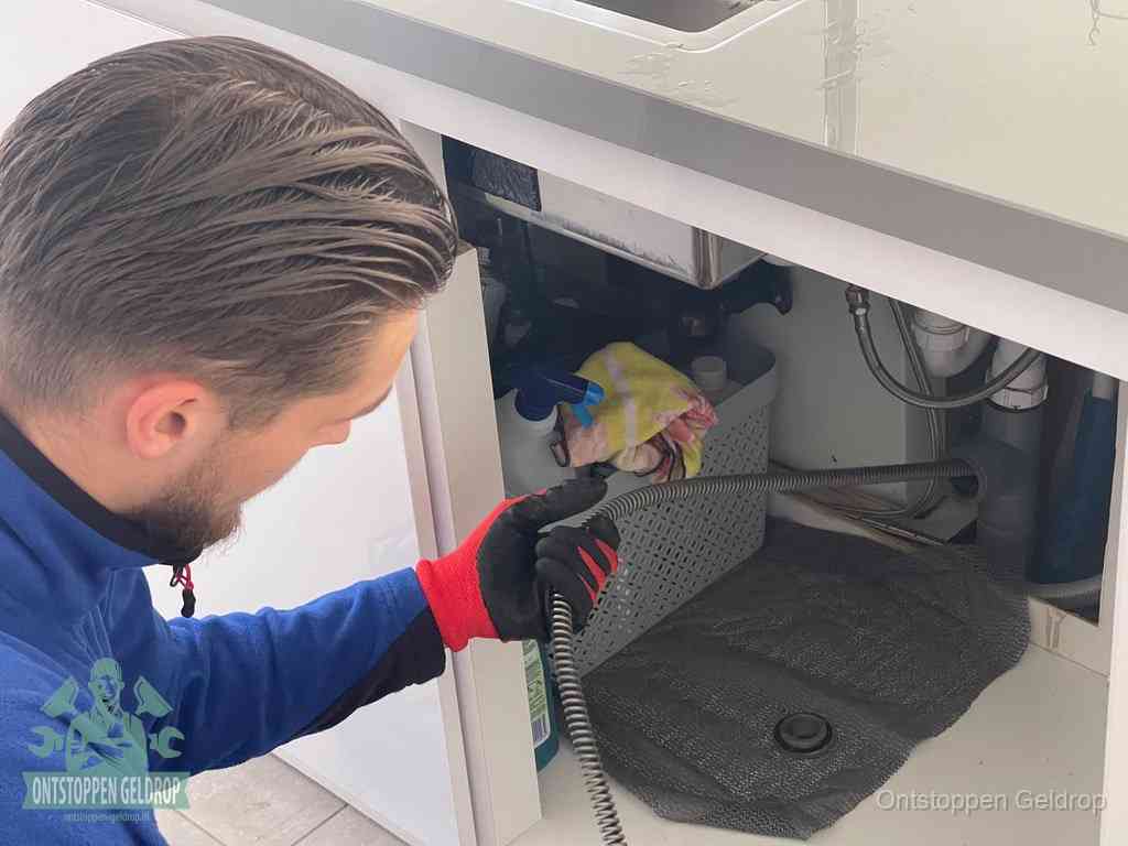 Een monteur voert een flexibele ontstoppingsveer in onder een keukenkastje om een verstopping voorbij de sifon te bereiken in Geldrop