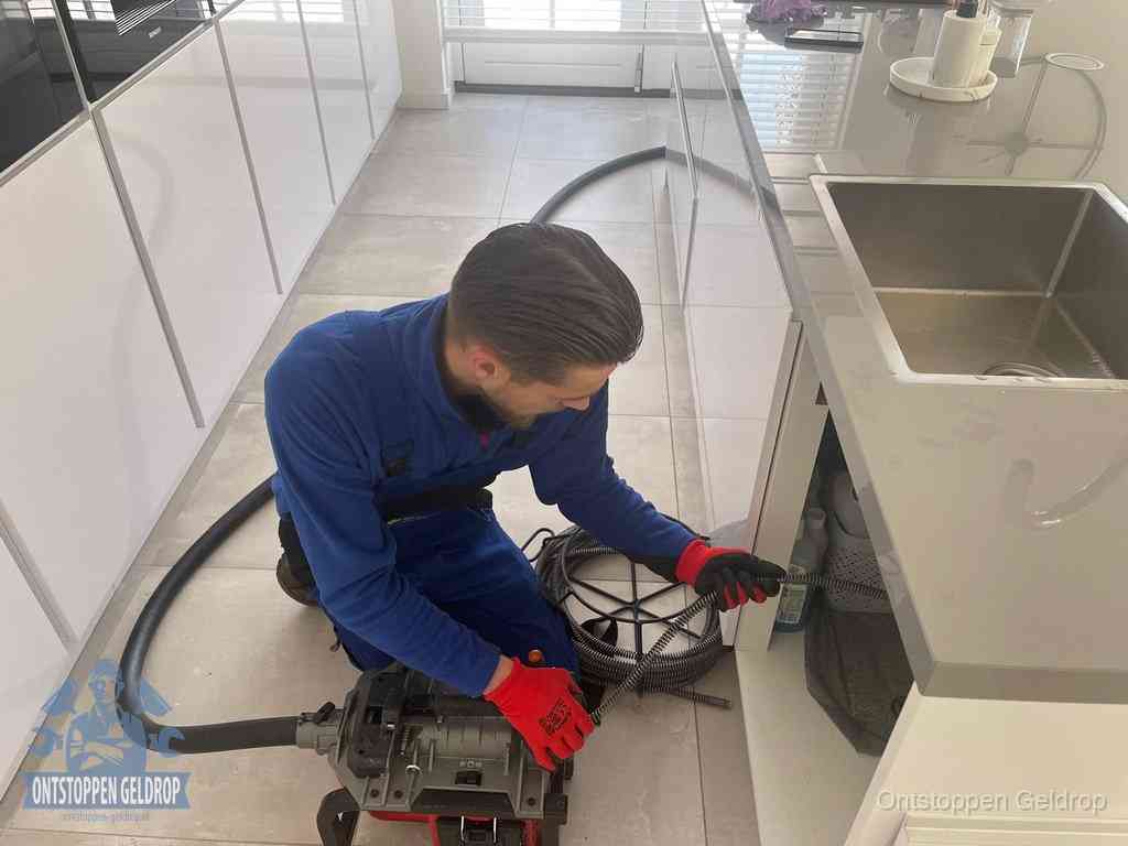 Een monteur ontstopt de keukenafvoer onder de spoelbak met een elektrische veermachine in Geldrop