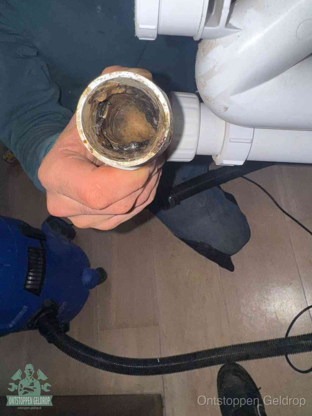 Een hand houdt een losgekoppeld PVC-afvoerbuisstuk vast dat van binnen volledig is dichtgeslibd met vet, vuil en aanslag, de meest voorkomende oorzaak van een verstopte afvoer in Geldrop