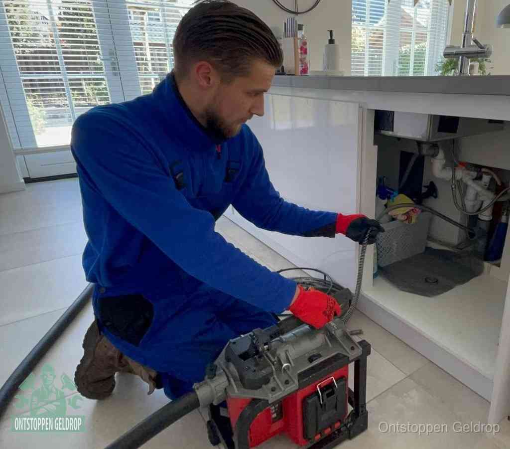 Een monteur met rode handschoenen knielt in een keuken en voert een elektrische ontstoppingsveer in tijdens het professioneel afvoer ontstoppen in Geldrop