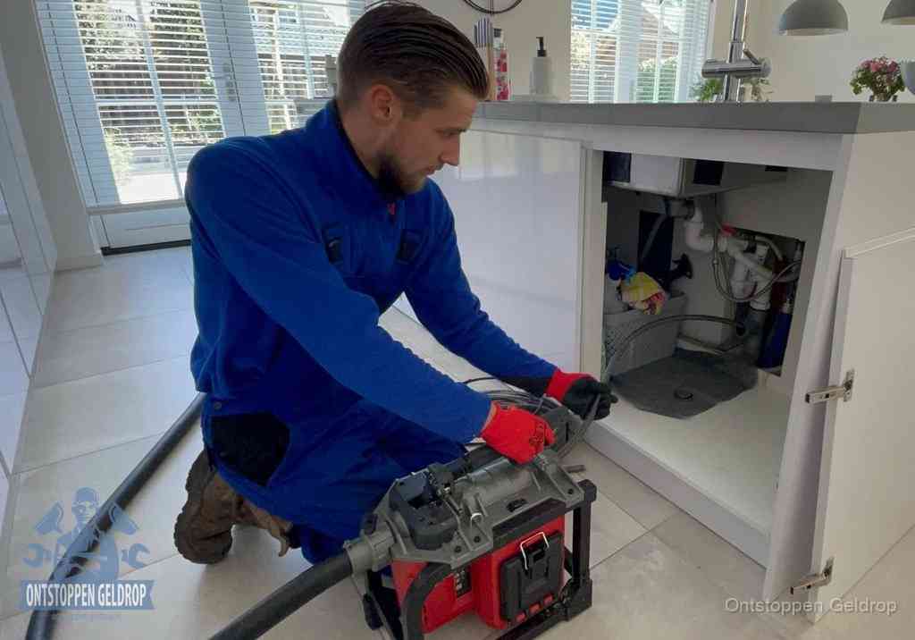 Een monteur voert een professionele elektrische ontstoppingsmachine in de afvoer onder de spoelbak tijdens het gootsteen ontstoppen in Geldrop